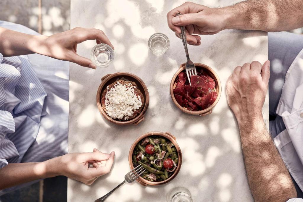 Divi cilvēki dalās maltītē, baudot dažādus ēdienus no zemām traukiem uz galda, apgaismojot ar dabiskiem ēnu rakstiem.