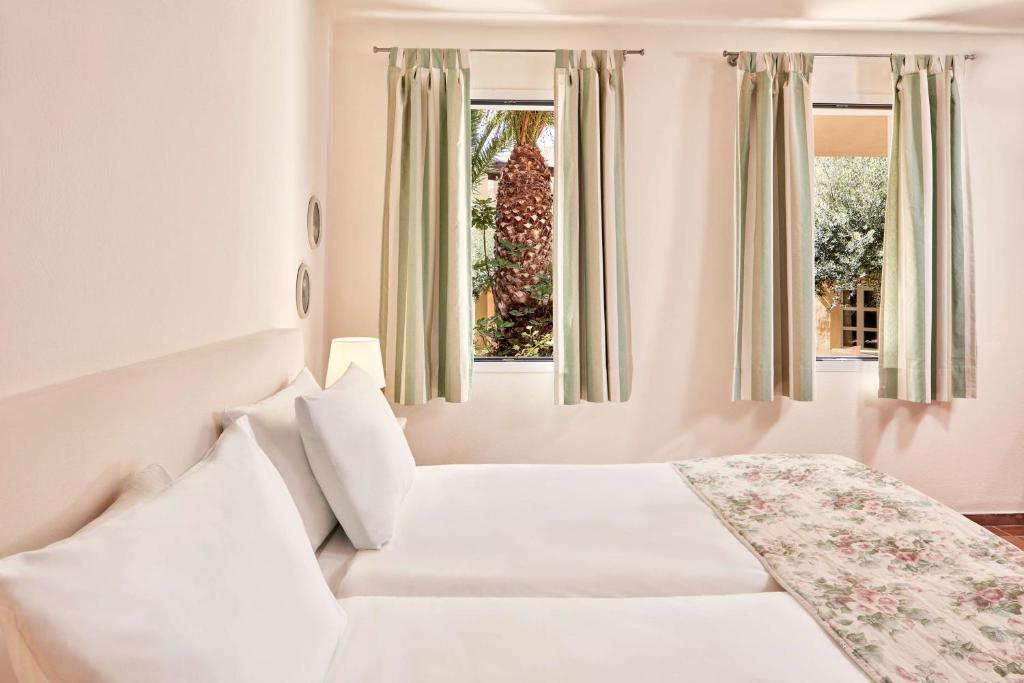 Gaiša guļamistaba ar divām gultām, logiem ar zaļām svītrainām aizkariem un skatu uz dārzu. Interjers ir vienkāršs un mājīgs, radot relaksējošu atmosfēru.