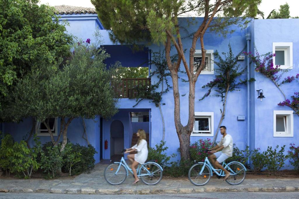 Divi cilvēki brauc ar velosipēdiem gar zilu māju, ko ieskauj zaļi koki un ziedi. Mājas fasāde ir dekorēta ar baltiem logiem un dzīvīgo veģetāciju.