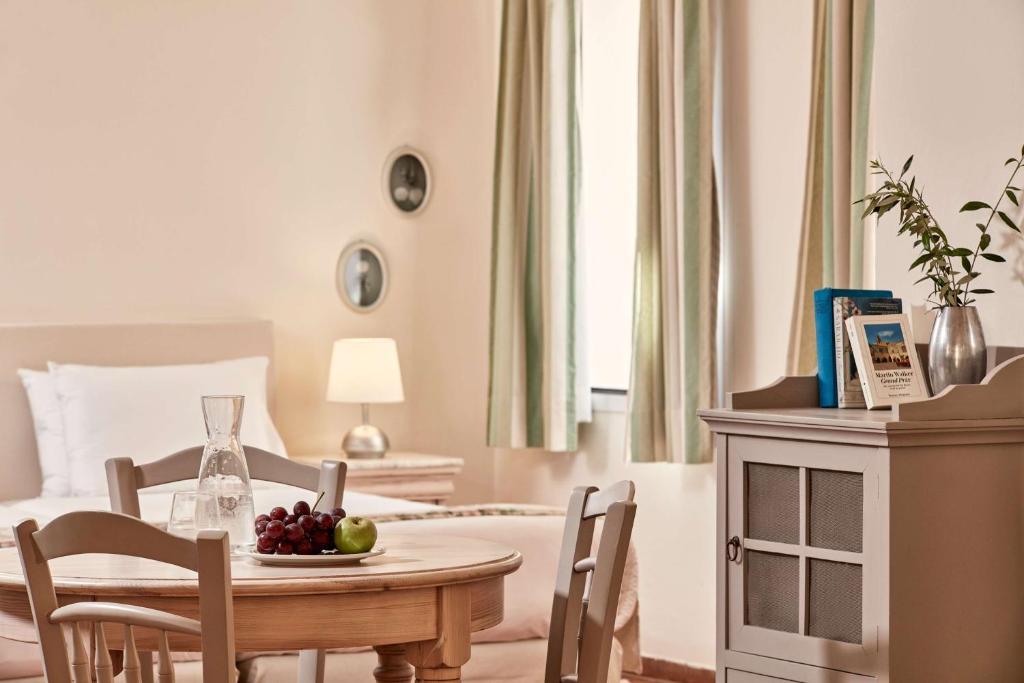 Gaiša un mājīga viesnīcas istaba ar koka galdu, kurā ir augļi un ūdens karafe. Fons ietver gultu ar baltām gultasveļām un logus ar gaišām aizkariem.