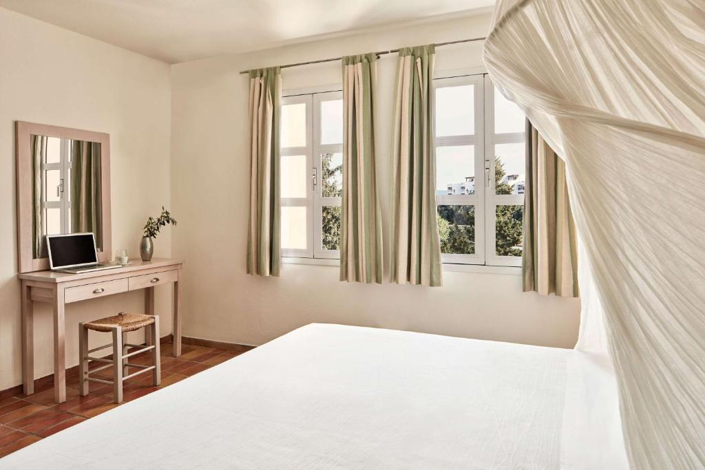 Mūsdienīga guļamistaba ar gaisīgu interjeru, logiem un koka mēbelēm, kas rada mierīgu atmosfēru. Gultā ir balts pārklājs un blakus novietota rakstāmgalds ar datoru.