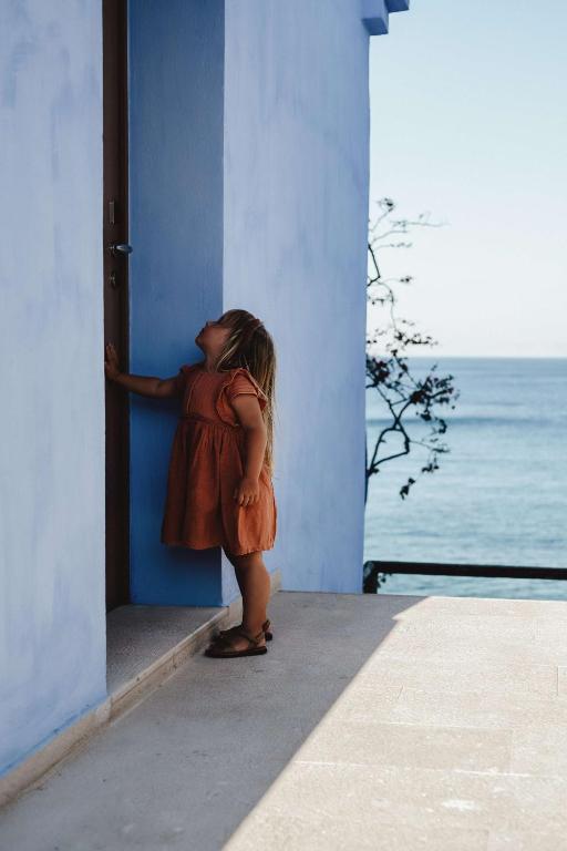 Bērns oranžā kleitā stāv pie zila mājas durvīm, skatoties uz augšu. Fons rāda jūru un debesis, radot mierīgu un idillisku atmosfēru.