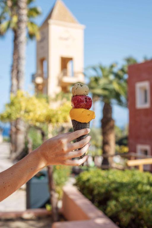 Krāsains saldējums uz vafeles konusa, turēts rokā, ar palmām un ēku fona. Ideāls vasaras deserts.