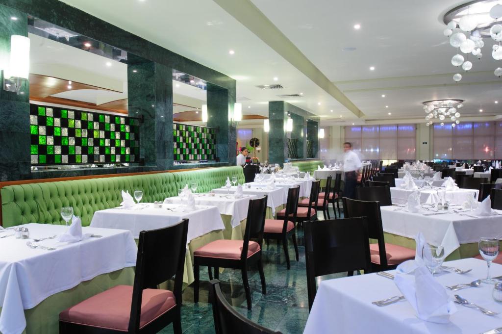 Elegants restorāns ar zaļām dīvāna sēdvietām un baltām galdautām. Interjerā izceļas mūsdienīgas lampas un dekoratīvi elementi, radot patīkamu atmosfēru.