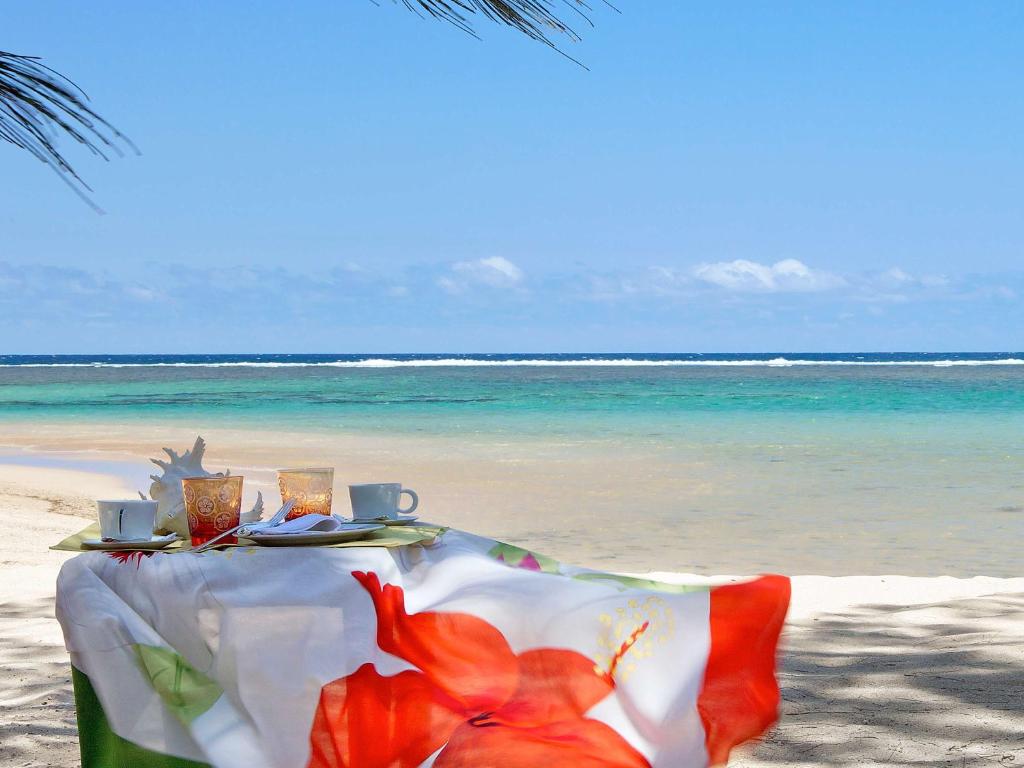 Pludmales galds ar brokastīm, dekorēts ar ziedu rakstu, ar skatu uz skaidru zilo jūru un smiltīm. Ideāla vieta relaksācijai un atpūtai.