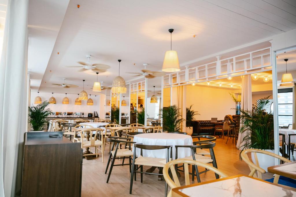 Mūsdienīgs restorāna interjers ar gaišām krāsām, koka mēbelēm un rotājošām lampām, kas rada mājīgu atmosfēru. Redzamas zaļās augu kompozīcijas un plašs ēdamzāles izkārtojums.