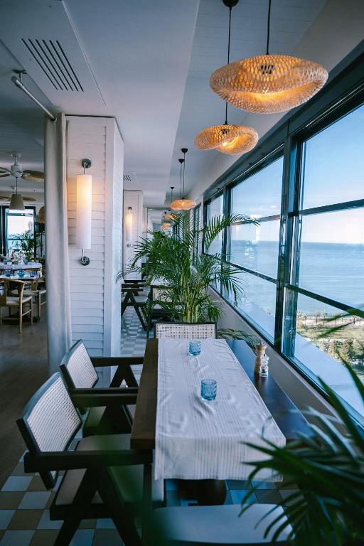 Mūsdienīgs restorāns ar gaišu interjeru, plašām logu rindām un skatu uz jūru, rotāts ar augiem un ērtām sēdvietām.