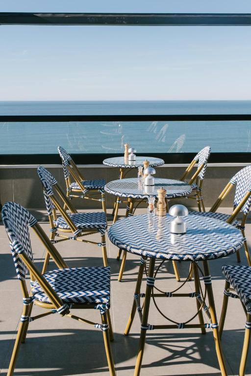 Mūsdienīgs kafejnīcas interjers ar zilu un balto rakstu galdiem un krēsliem, kas atrodas pie jūras. Skats uz okeānu rada relaksējošu atmosfēru.