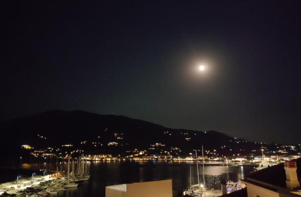 Mēness apgaismo nakts ainavu virs ostas, kur redzami jahti un gaismas kalnos. Klusa un burvīga atmosfēra.