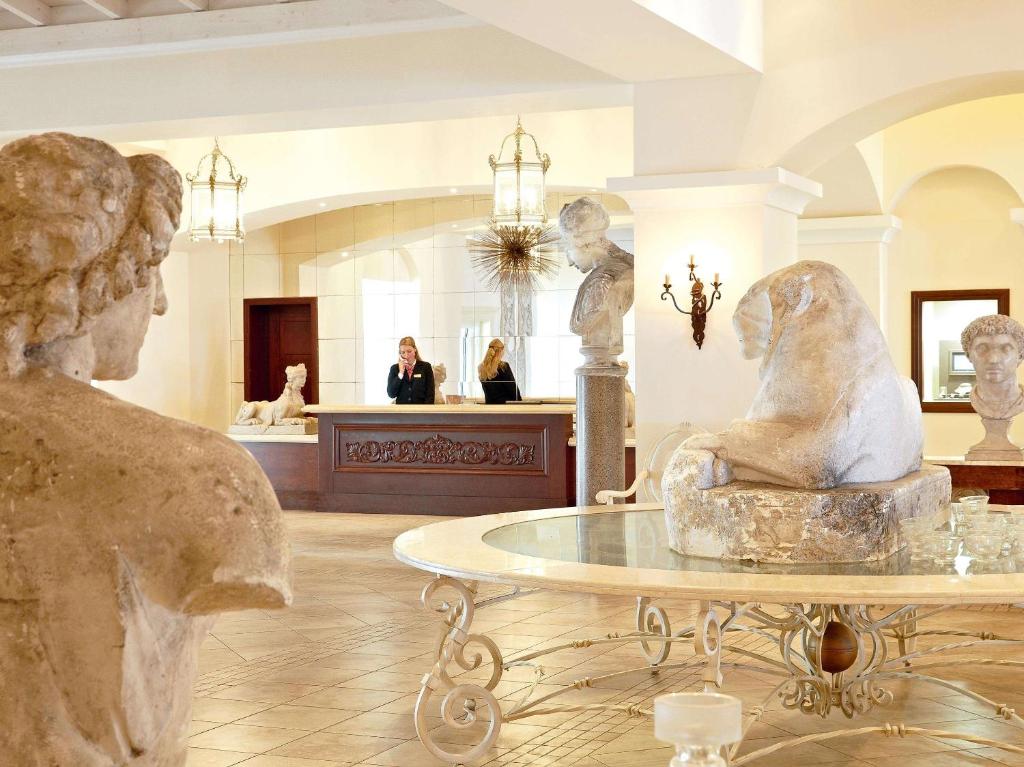 Elegants viesnīcas interjers ar klasiskām skulptūrām un stilu. Reģistratūras apkalpojošais personāls ir redzams fonā, radot viesmīlīgu atmosfēru.