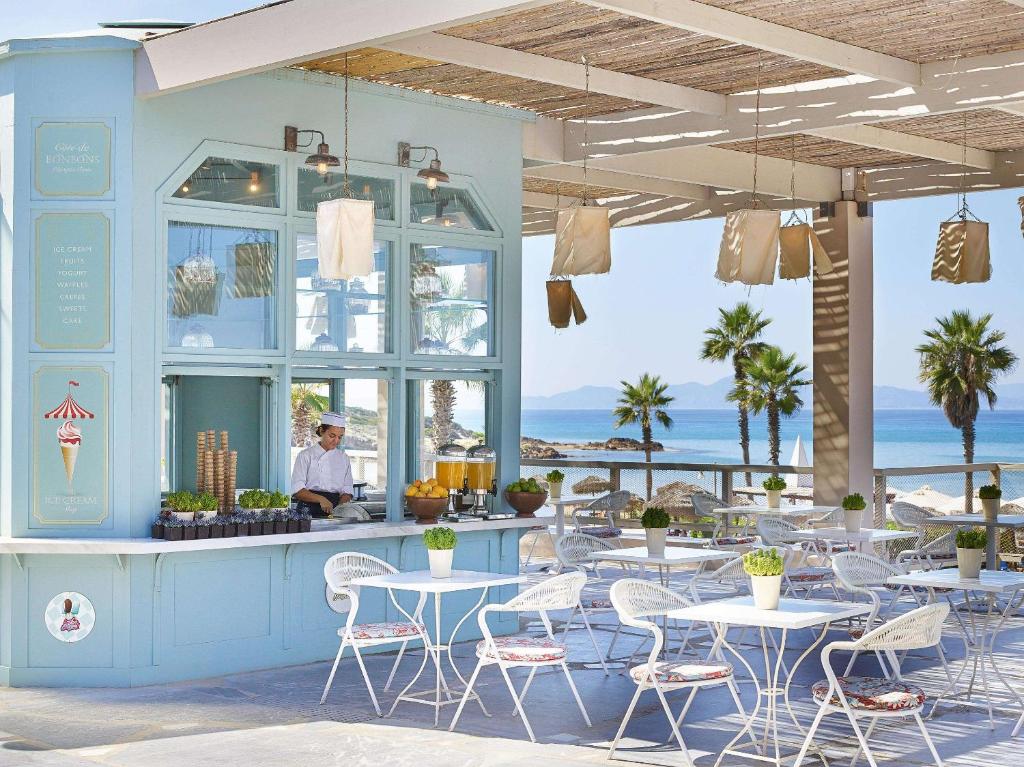 Pludmales kafejnīca ar gaišu interjeru, kurā redzams viesmīlis, gatavojot ēdienus. Apkārt ir balti galdi un krēsli, kā arī palmas un jūras skats.