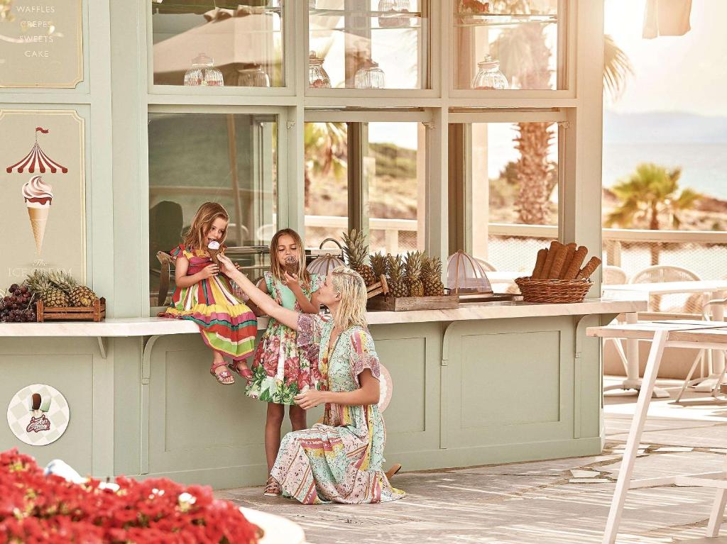 Trīs meitenes, no kurām viena sēž uz letes un ēd saldējumu, bet otra pieaugušā sieviete, tērpta krāsainā apģērbā, ar prieku sniedz viņai saldējumu. Fons ir gaišs un saulains, ar palmām un pludmales skatu.