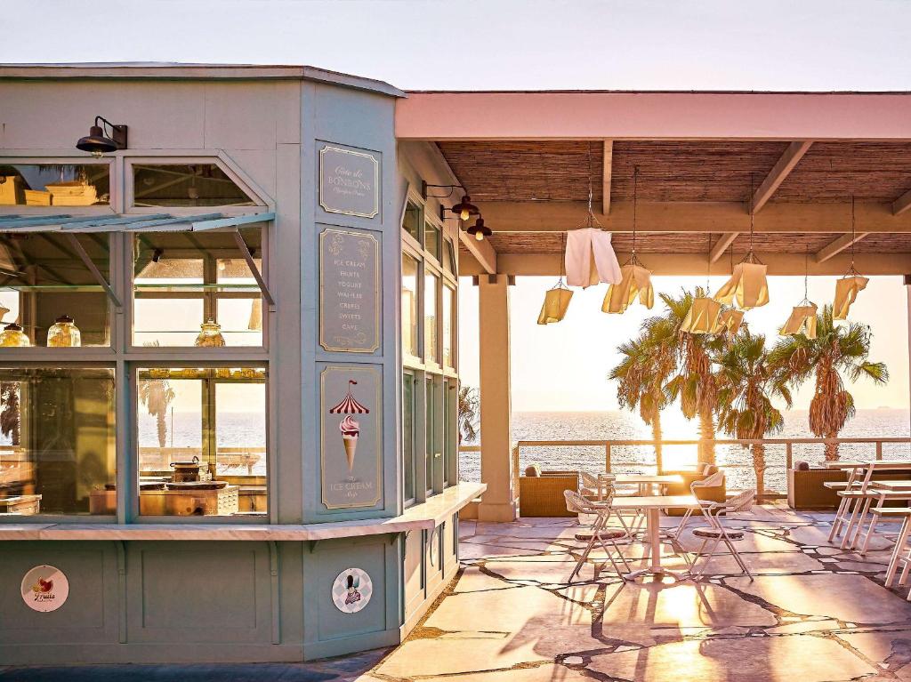 Kafejnīca pie jūras ar gaišu interjeru un palmām, kas apņem terasi, kur saulriets rada siltu atmosfēru. Ideāla vieta atpūtai un baudīšanai.