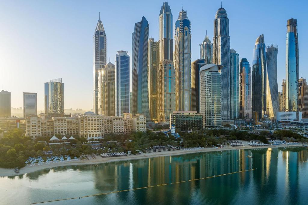 Dubaijas pilsētas ainava ar augstceltnēm un jūras krastu, atspoguļojoties ūdenī. Skats uz modernu arhitektūru un saulrieta gaismu.