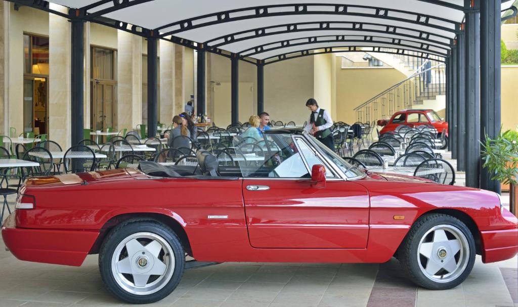 Sarkans kabriolets atrodas modernā terasē ar ēdināšanas zonu, kur apmeklētāji sēž pie galdiem. Fons rāda stilīgu arhitektūru un citus automobiļus.