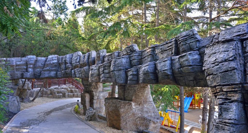Aizraujoša akmens tilta struktūra virs pastaigu celiņa, ko ieskauj bagātīga zaļā daba un bērnu spēļu laukums.