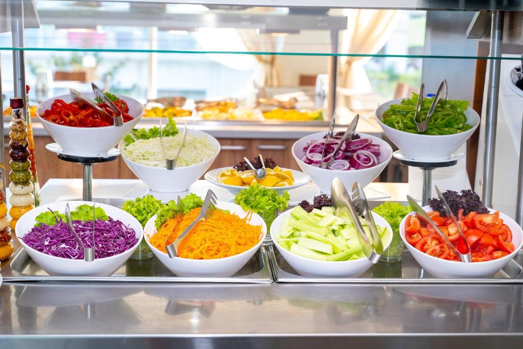 Svaigu dārzeņu bufete ar dažādām salātu sastāvdaļām, tostarp burkāniem, tomātiem, kāpostiem un paprikām. Krāsaini dārzeņi ir izkārtoti baltos traukos, radot pievilcīgu un veselīgu ēdienu piedāvājumu.