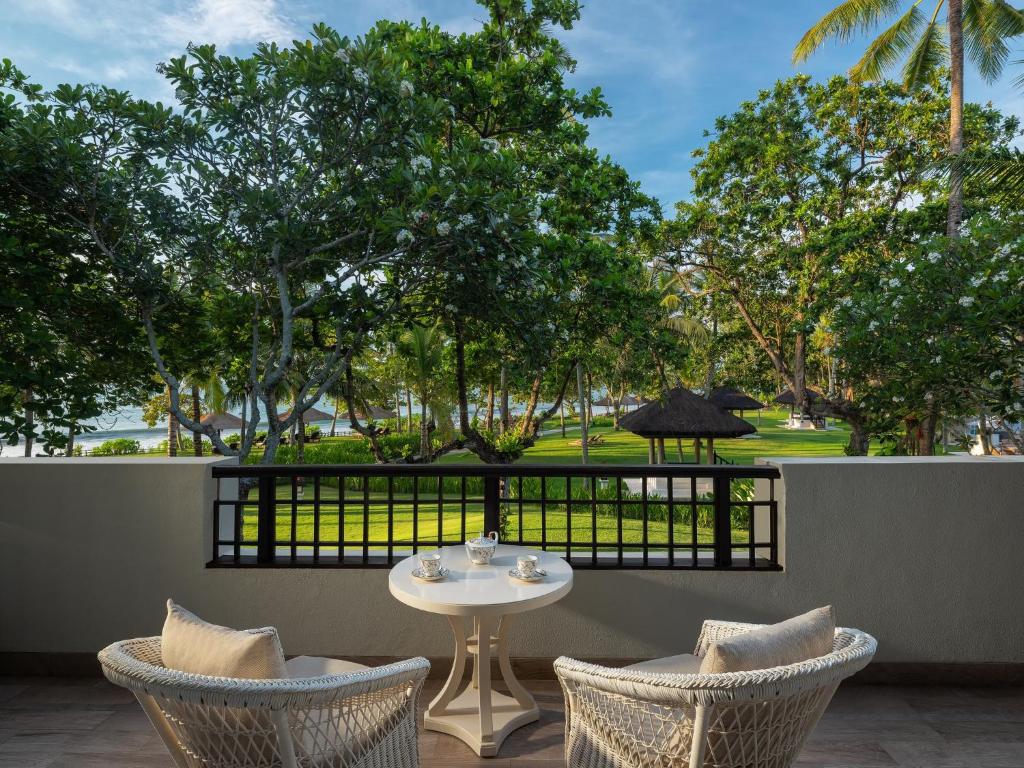 Balkons ar divām krēslām un galdu, no kura paveras skats uz zaļu dārzu un tuvumā esošo pludmali. Apkārt aug koki un palmas, radot mierīgu un relaksējošu atmosfēru.