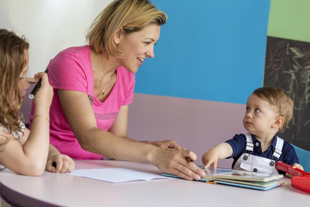 Mamma strādā ar bērniem, palīdzot viņiem mācīties, kamēr viņi ir sēdējuši pie galda. Gaisīgā un krāsainā telpā redzami divi mazi bērni, no kuriem viens uzmanīgi skatās uz grāmatu.