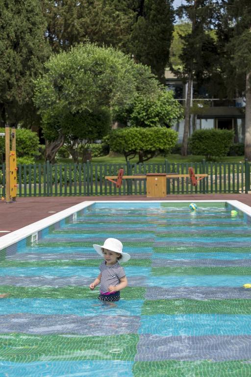 Bērns ar baltu cepuri rotaļājas seklajā baseinā, apkārt ir zaļi koki un labi kopts parks. Baseins ir izrotāts ar krāsainu mozaīku.