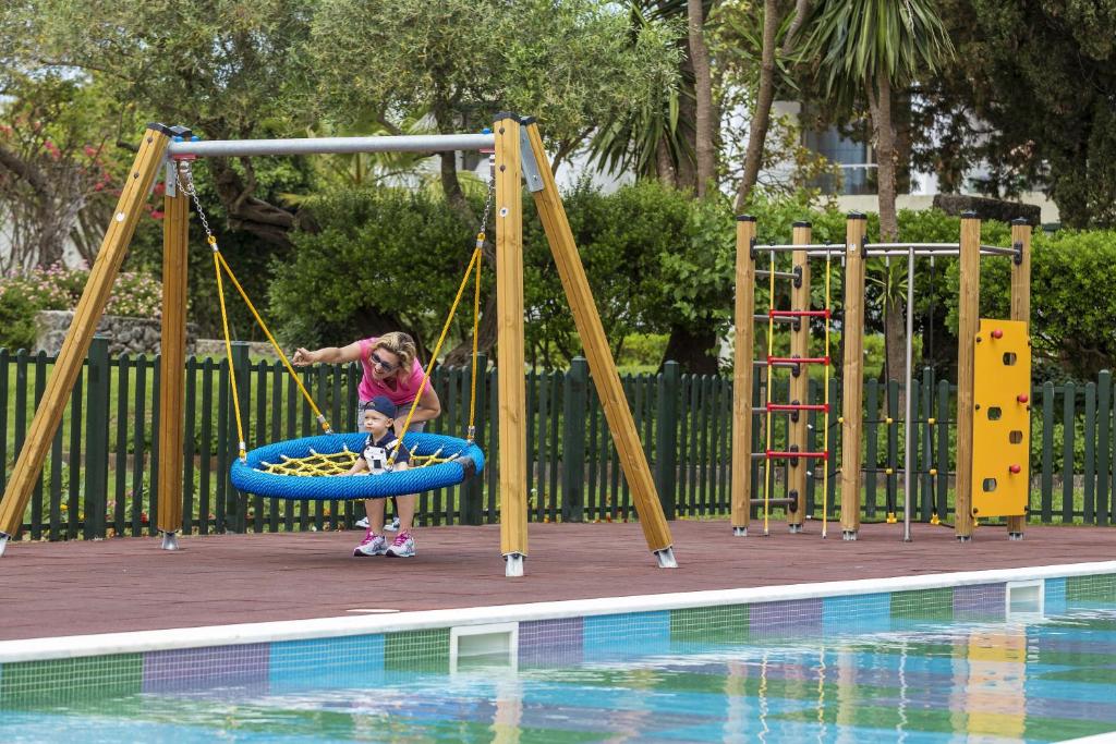 Bērnu rotaļu laukums ar šūpolēm un kāpšanas sienu, kur māte palīdz bērnam spēlēties. Apkārt ir zaļi krūmi un dīķis.