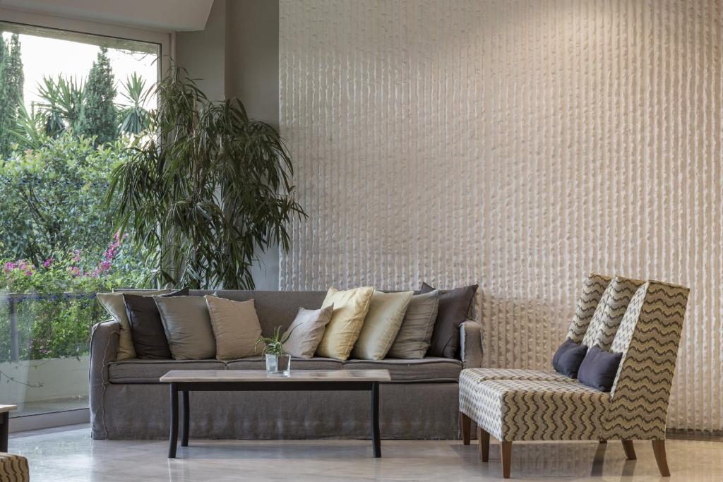 Mūsdienīgs dzīvojamās istabas interjers ar dīvānu, spilveniem un rakstainu krēslu. Loga blakus redzami zaļumi un ziedi, radot patīkamu atmosfēru.