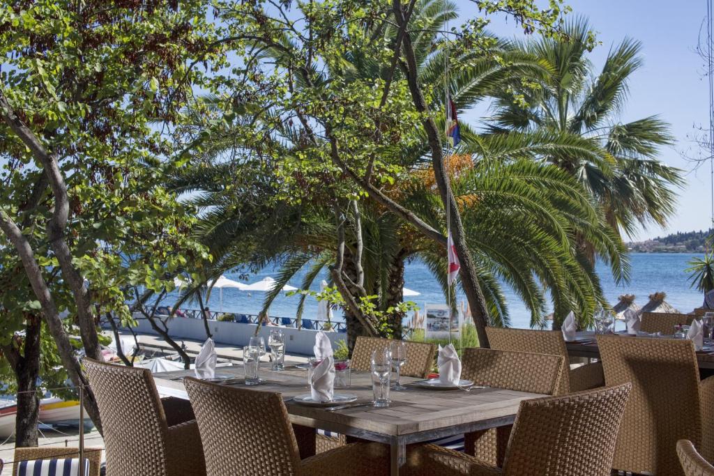 Restorāna terase ar skatu uz jūru, apzaļumotu ar palmām un lapotnēm, radot relaksējošu atmosfēru. Galdi ir sakārtoti ar galdautiem un traukiem, gatavi viesu sagaidīšanai.
