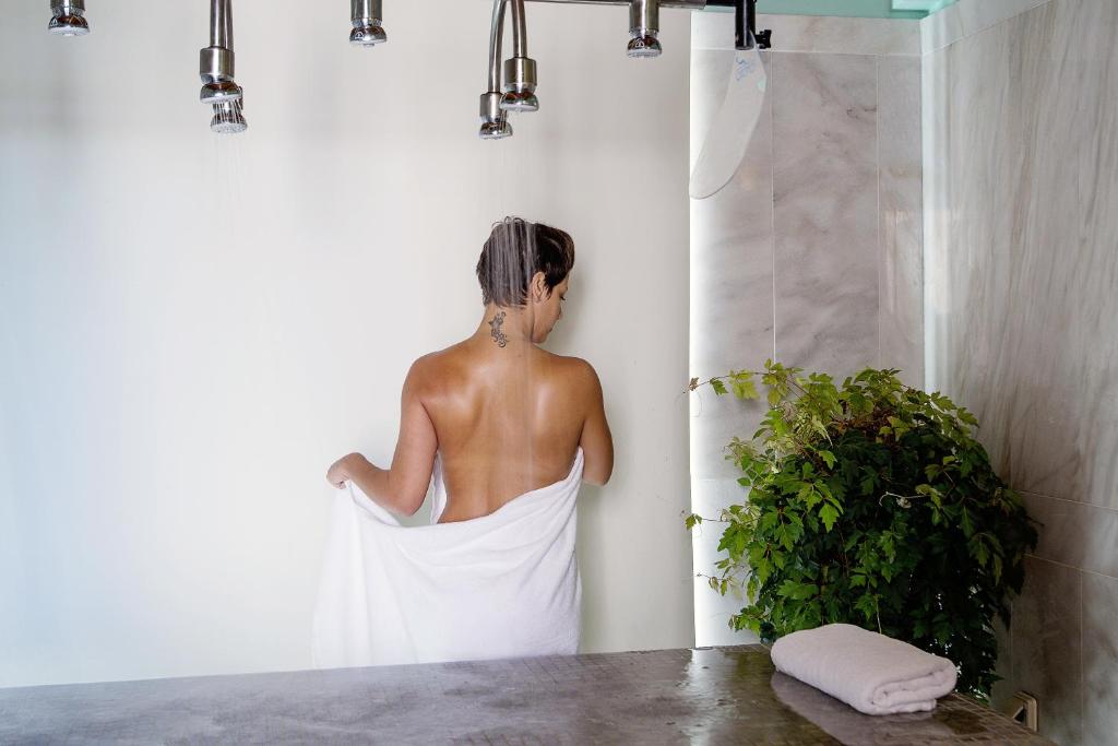 Sieviete ar īsiem matiem stāv dušā, apņemta ar baltu dvieli. Fons ir mūsdienīga vannas istaba ar dabīgām zaļām augu detaļām.