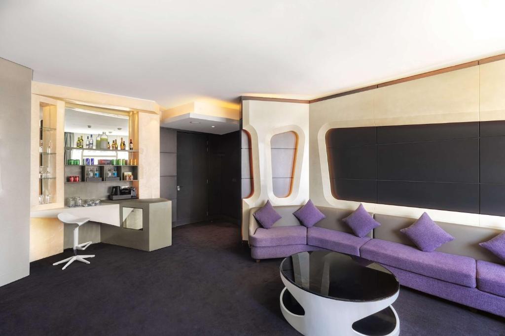 Mūsdienīgs viesnīcas numurs ar ērtu purpura krāsas dīvānu un modernu bāra zonu. Interjers apvieno eleganci un komfortu, radot patīkamu atmosfēru.