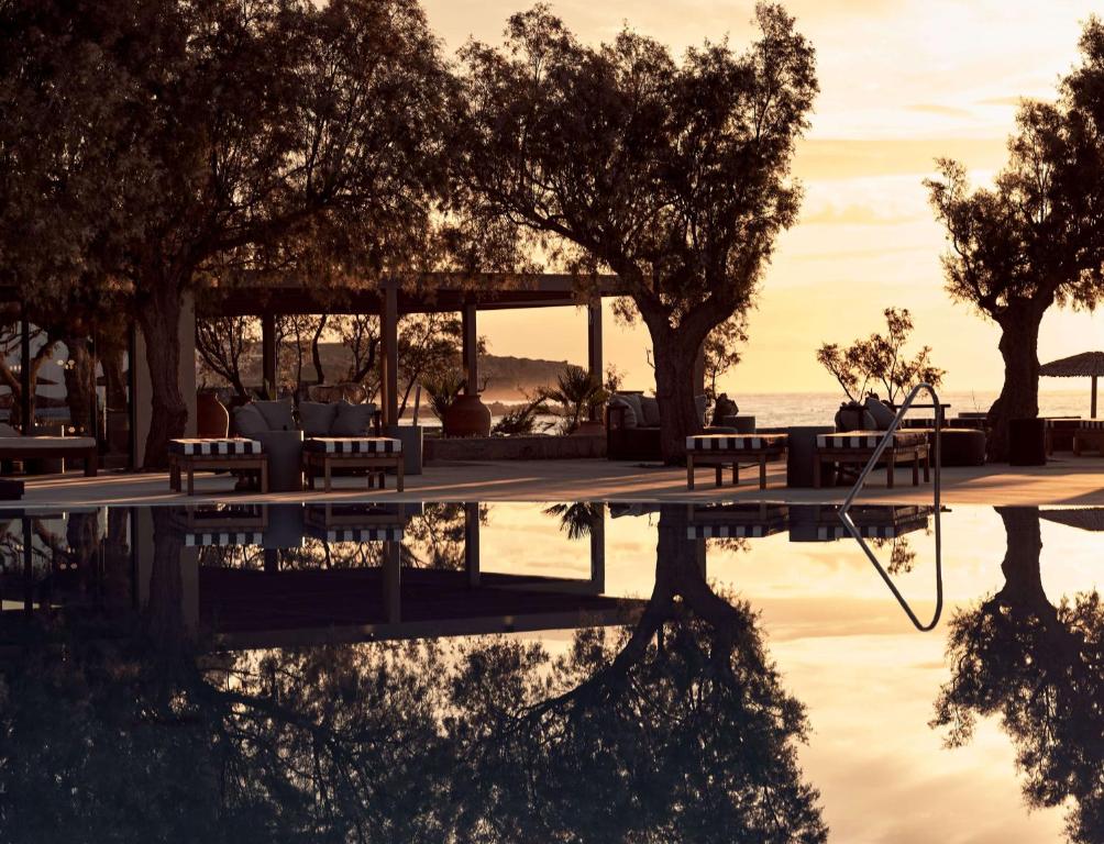 Elegants baseins ar atspulgu, apkārt ir koka terase un ērtas atpūtas zonas, zem kokiem. Fons veido saulriets pie jūras, radot mierīgu atmosfēru.