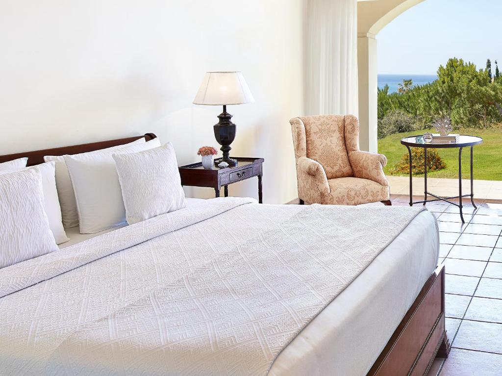 Mūsdienīgs guļamistabas interjers ar plašu gultu, mīkstām spilveniem un ērtiem krēsliem, no kura paveras skats uz dārzu un jūru.