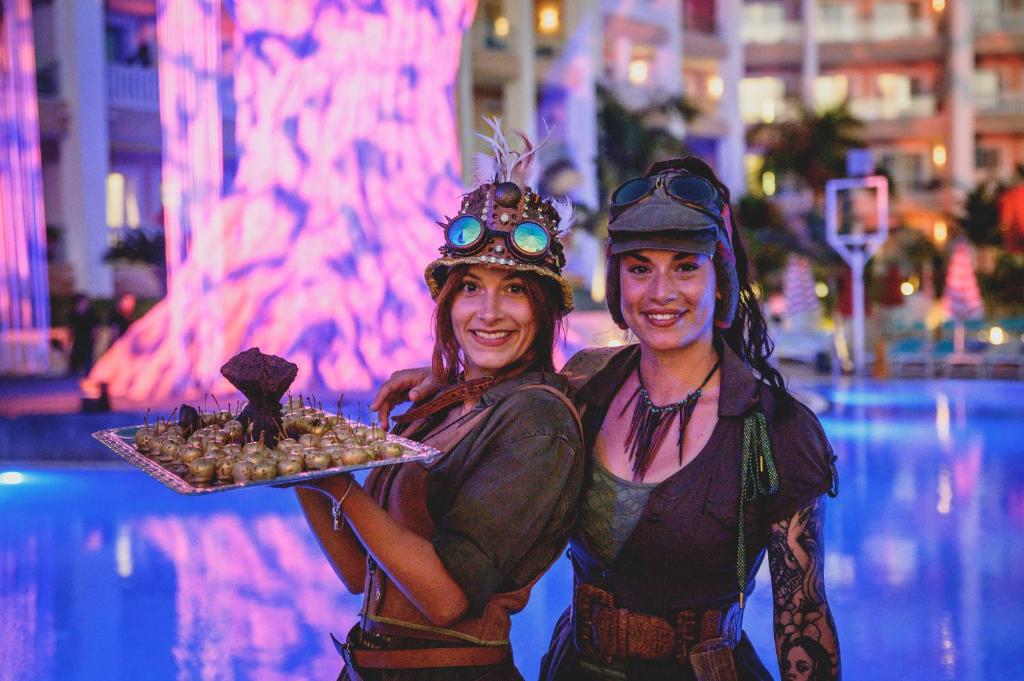 Divas sievietes tērptas steampunk stilā, tur rokās paplāti ar izsmalcinātiem uzkodu ēdieniem, fona apgaismojumā redzama krāsaina gaismas instalācija.