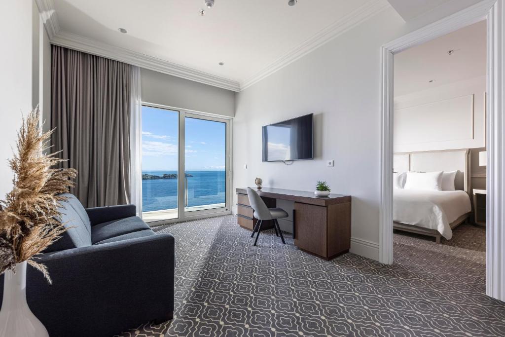 Mūsdienīgs viesnīcas numurs ar skatu uz jūru, kurā ir dīvāns, rakstāmgalds un televizors. Telpā ir gaišas krāsas un lieli logi, kas piepilda to ar dabiski gaismu.