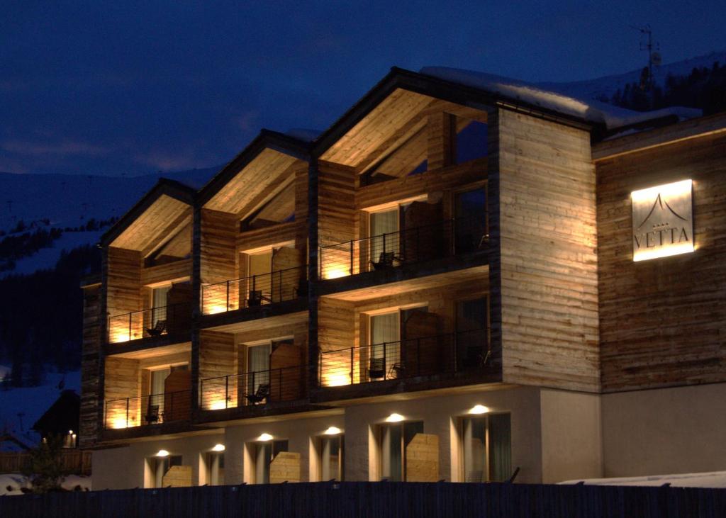 Mūsdienīgs kalnu viesnīcas eksterjers ar koka apdari un izgaismotiem balkoniem, kas redzams tumsā. Viesnīcas nosaukums "Vetta" ir redzams uz sienas.