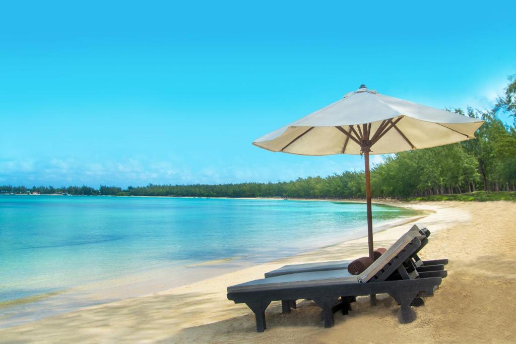 Pludmales ainava ar sauļošanās krēsliem un lietussargu, kas atrodas pie mierīga ūdens, ar zilu debesu fonu. Ideāla vieta relaksācijai un atpūtai.