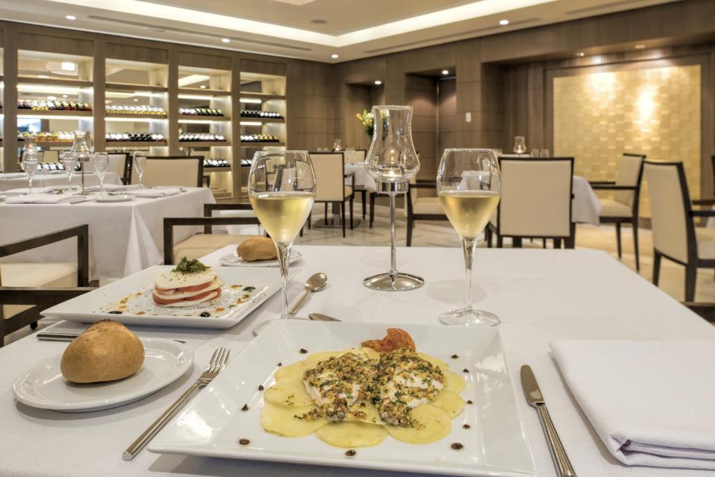Elegants restorāns ar izsmalcinātu galda klājumu, uz kura redzama garšīga maltīte un glāze baltvīna. Fons izrotāts ar vīna plauktiem un modernu interjeru.