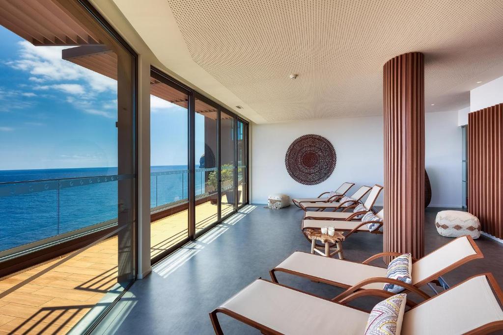 Mūsdienīgs spa telpas interjers ar lieliem logiem, kas sniedz skatu uz jūru, un komfortablām atpūtas krēslām. Zālē ir gaiša atmosfēra un eleganta dizaina elementi.