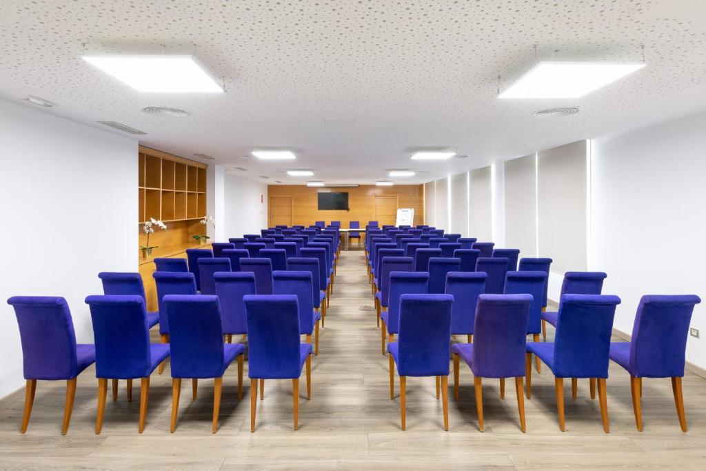 Konferenču telpa ar violetiem krēsliem, sakārtotiem rindās, un modernu interjeru. Telpā ir ekrāns un dabiskā gaisma, radot profesionālu atmosfēru.