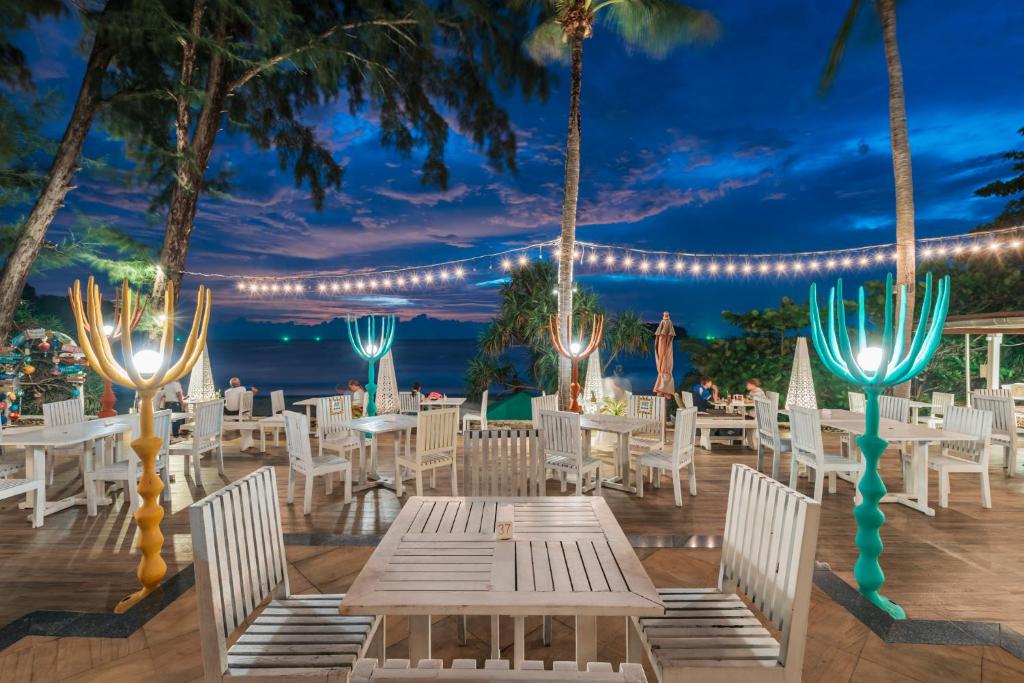 Lieliska pludmales restorāna terase ar koka galdiem un krēsliem, dekorēta ar stilizētām lampām un gaismas virtenēm, kas izgaismo vakara ainavu. Aiz terases redzams jūras skats un zilo debesu krāsas.