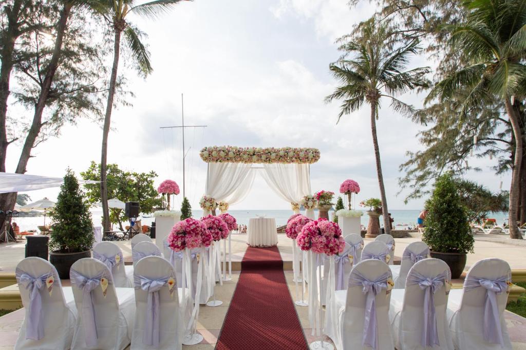 Romantiska pludmales kāzu ceremonija ar rozā ziediem, baltu altāri un dekoratīvām sēdvietām, kas izvietotas blakus palmām un jūrai.
