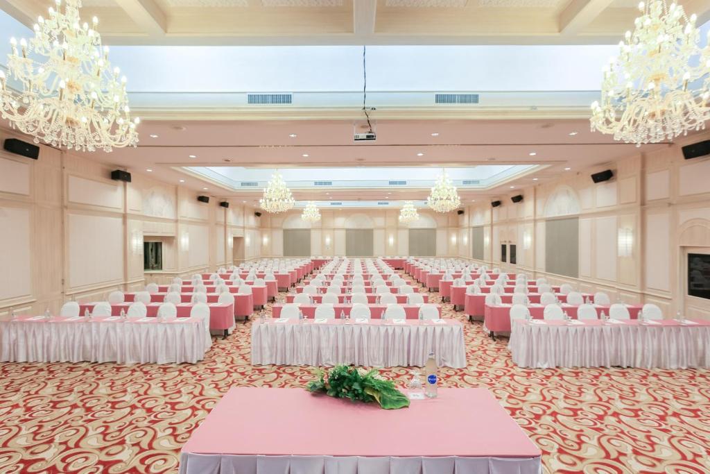 Liela un eleganta viesību zāle ar rozā galdautiem un baltām krēslām, kas ir sakārtoti rindās. Gaismas lustras piekārti pie griestiem, radot gaisīgu atmosfēru.