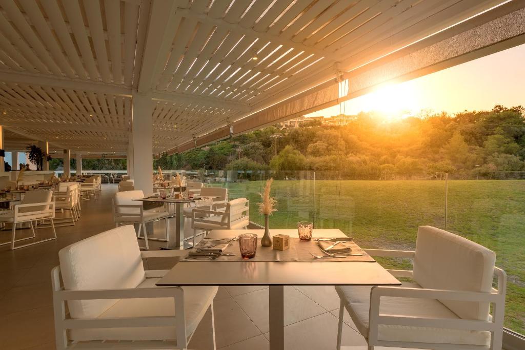 Mūsdienīgs restorāns ar baltām mēbelēm un panorāmas skatu uz saulrietu, kas izgaismo apkārtējo dabu. Dziļi relaksējoša atmosfēra, ideāla maltītei.