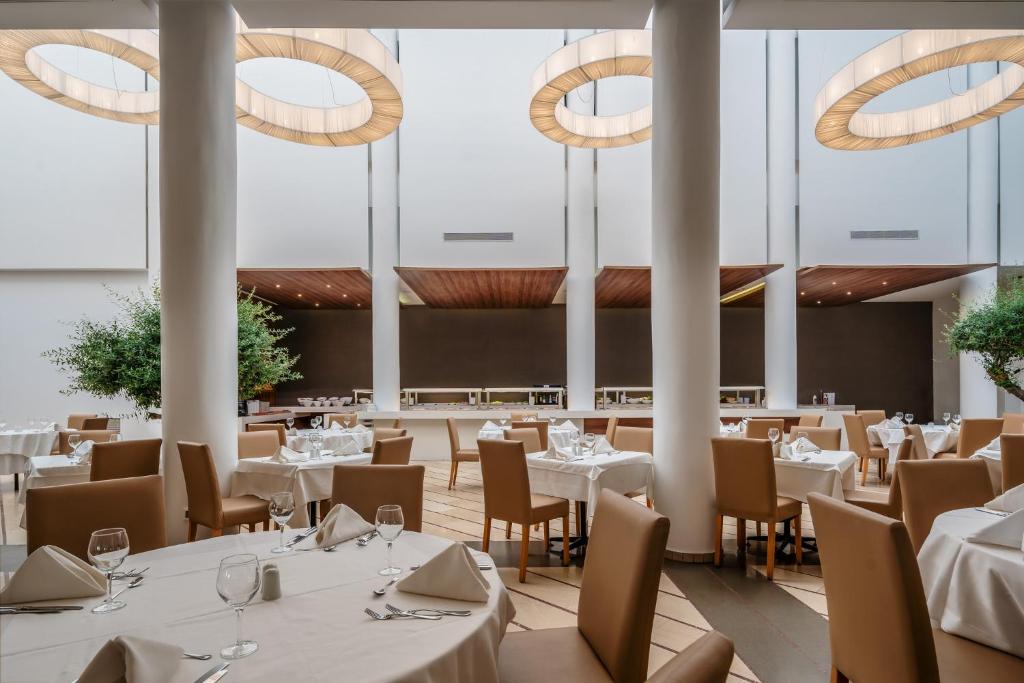 Mūsdienīgs restorāns ar elegantiem galdiem, baltiem galdautiem un stilīgām gaismas ķermeņiem. Interjers izceļas ar minimālisma dizainu un zaļiem augiem, radot patīkamu atmosfēru.