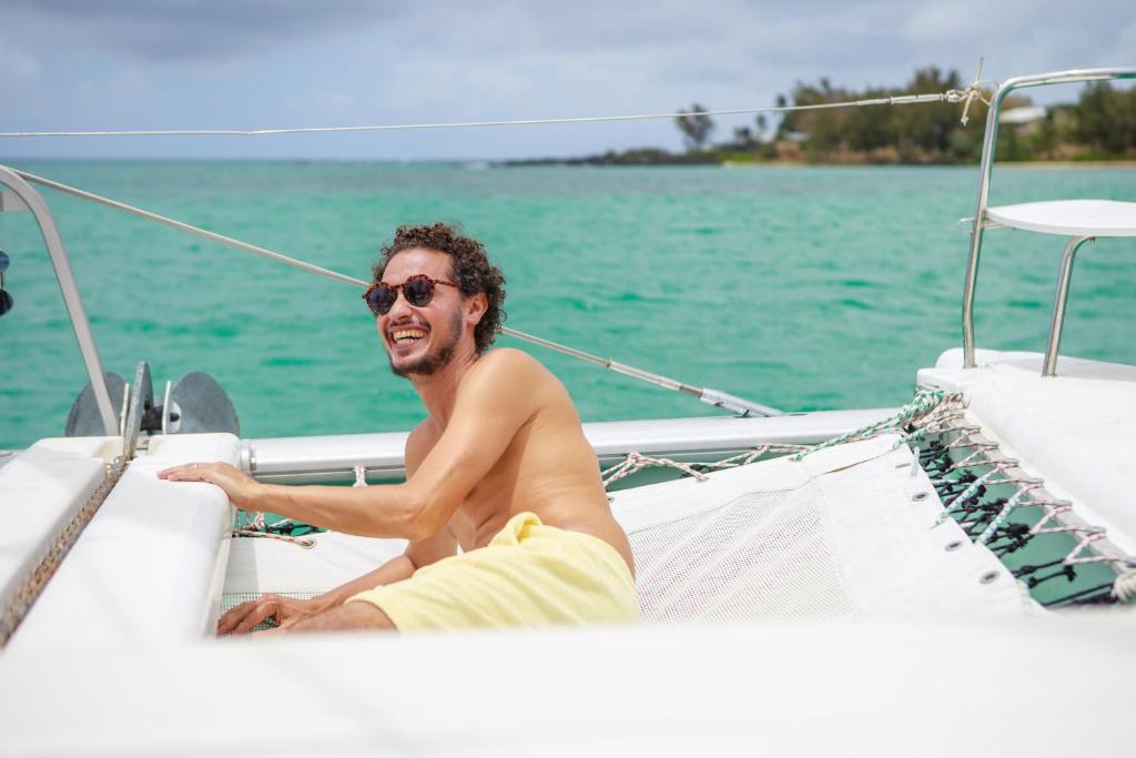 Jauns vīrietis ar saulesbrillēm atpūšas uz katamarāna, baudot sauli un jūras skatus. Apkārt ir skaista, zilza jūra un zaļi krasti.