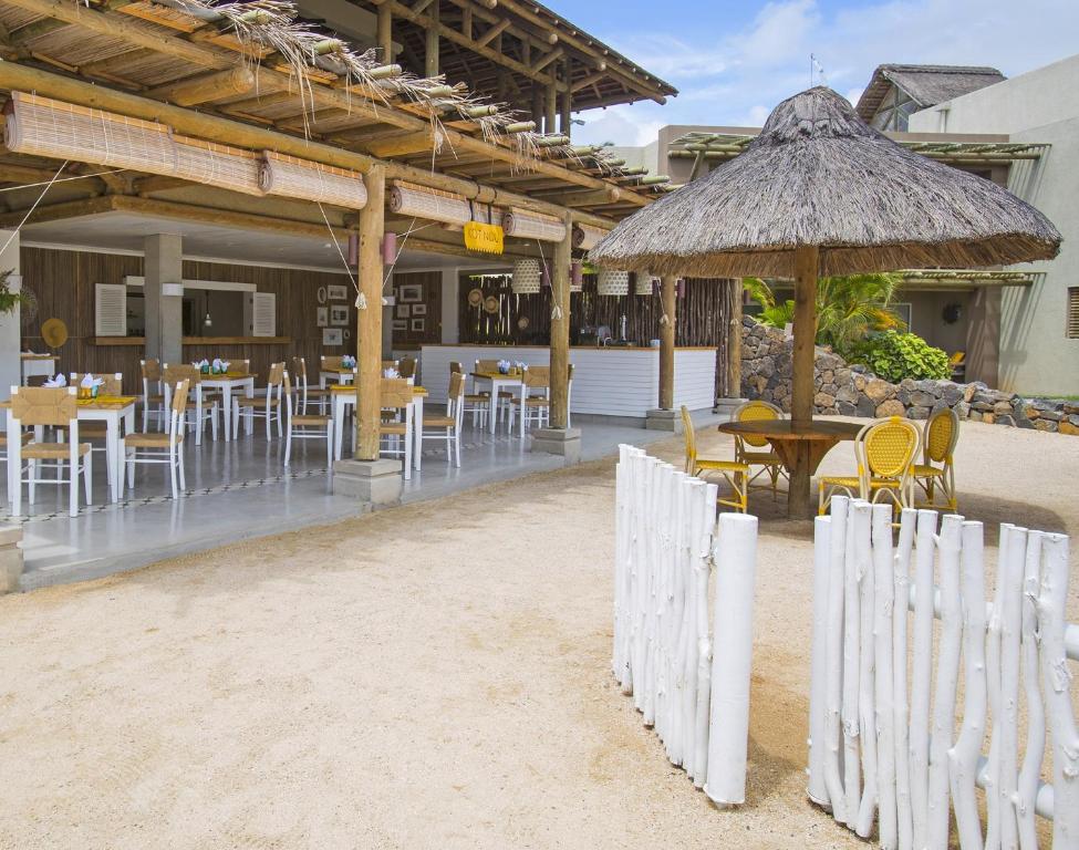 Brīvdabas restorāns ar tradicionālu jumtu un vieglu atmosfēru, kur redzami koka galdi un krēsli, kas izvietoti uz smiltīm. Ievērojama ir arī baltā žoga klātbūtne un apkārtējā zaļā ainava.