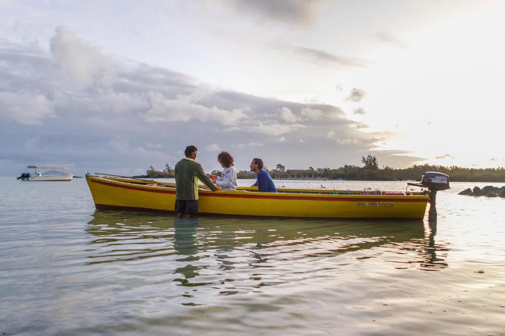 Trīs cilvēki jautri sarunājas laivā, kas stāv ūdenī, ar saulrietu fona. Laiva ir dzeltena un redzams motors aizmugurē.