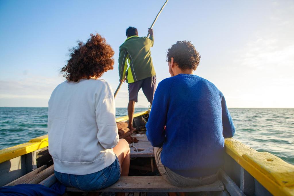 Divi cilvēki sēž laivā, skatoties uz jūru, kamēr laivas kapteinis airē. Gaisma ir silta un saulaina, radot relaksējošu atmosfēru.
