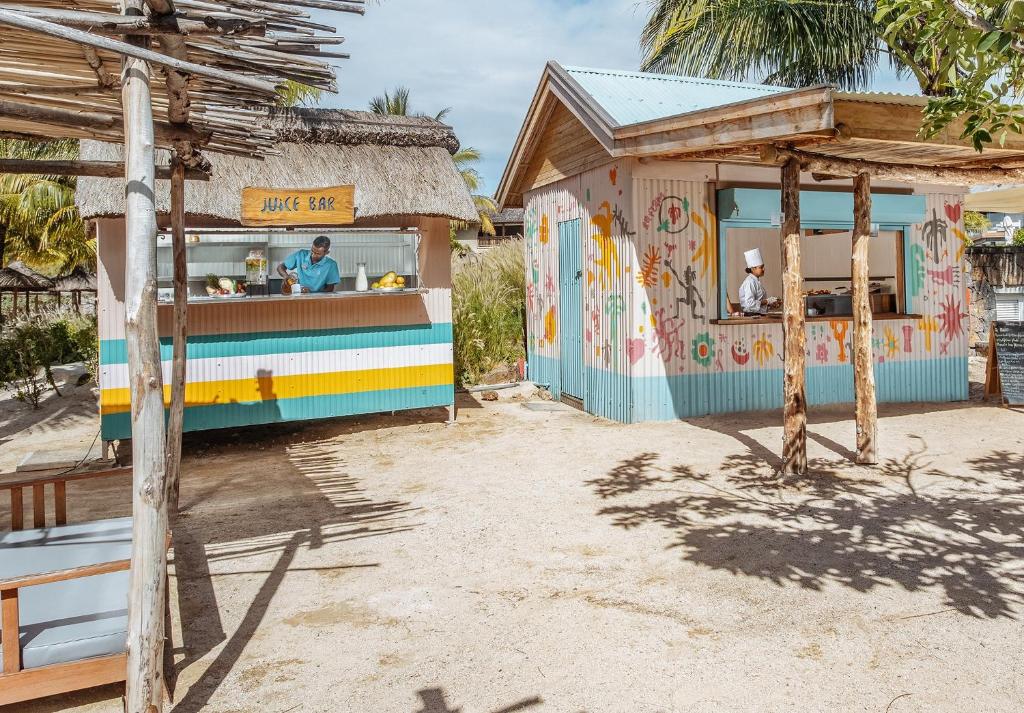 Krāsains augļu dzērienu bārs ar darbinieku, kas gatavo dzērienus, un krāsainā ēdienu stacija ar šefpavāru. Apkārt ir tropiskā vide ar palmām un smilšainu zemi.