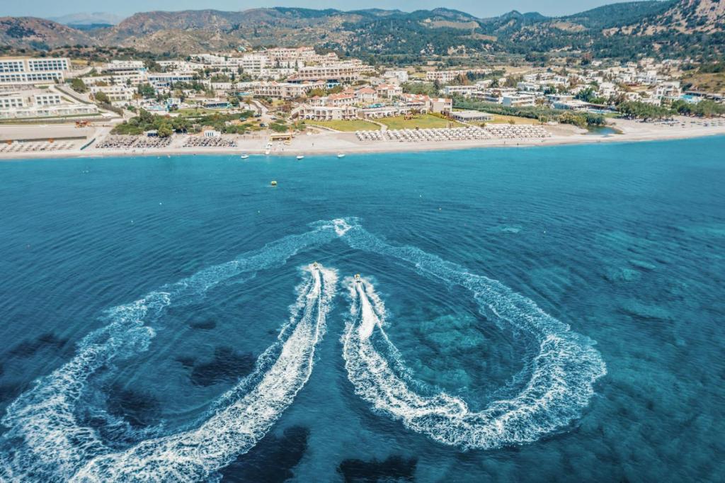 Skaista pludmales ainava ar skaidru zilu ūdeni un pludmali, kur redzami ūdens motocikli, kas atstāj viļņus. Fons ietver viesnīcas un kalnu ainavas.