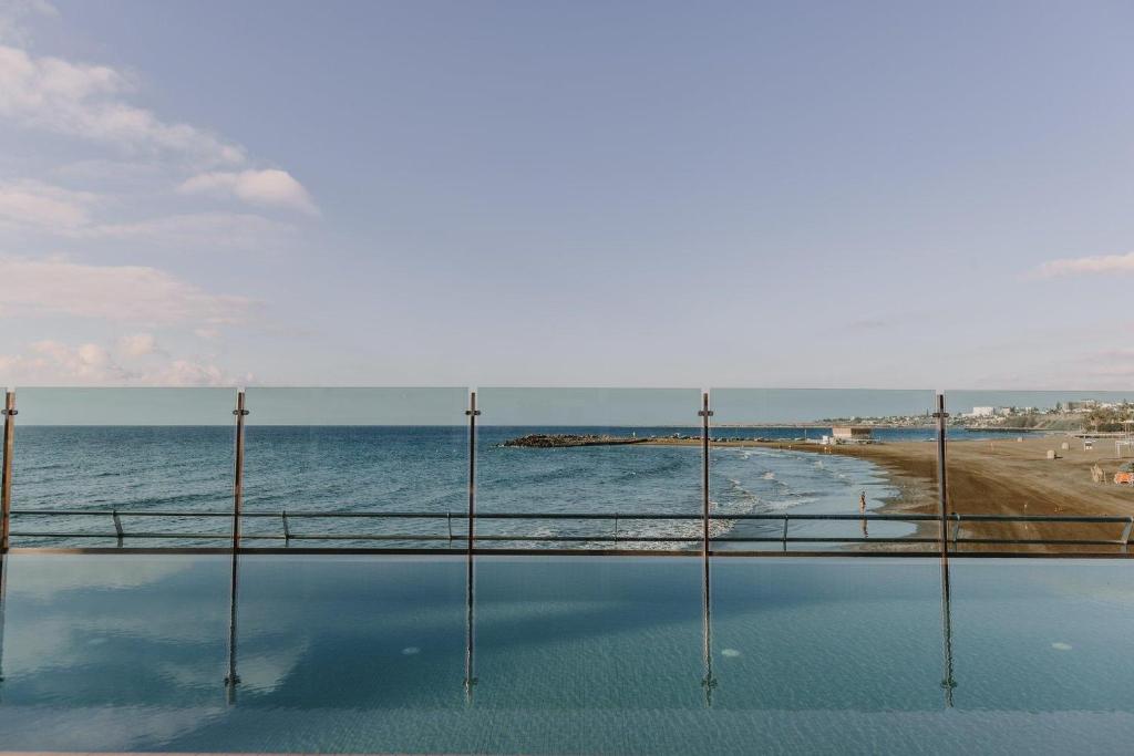 Skats uz mierīgu jūru ar pludmali un skaidru debesu fonu, ko norobežo stikla barjera. Ūdens virsma atspoguļo apkārtējo ainavu.